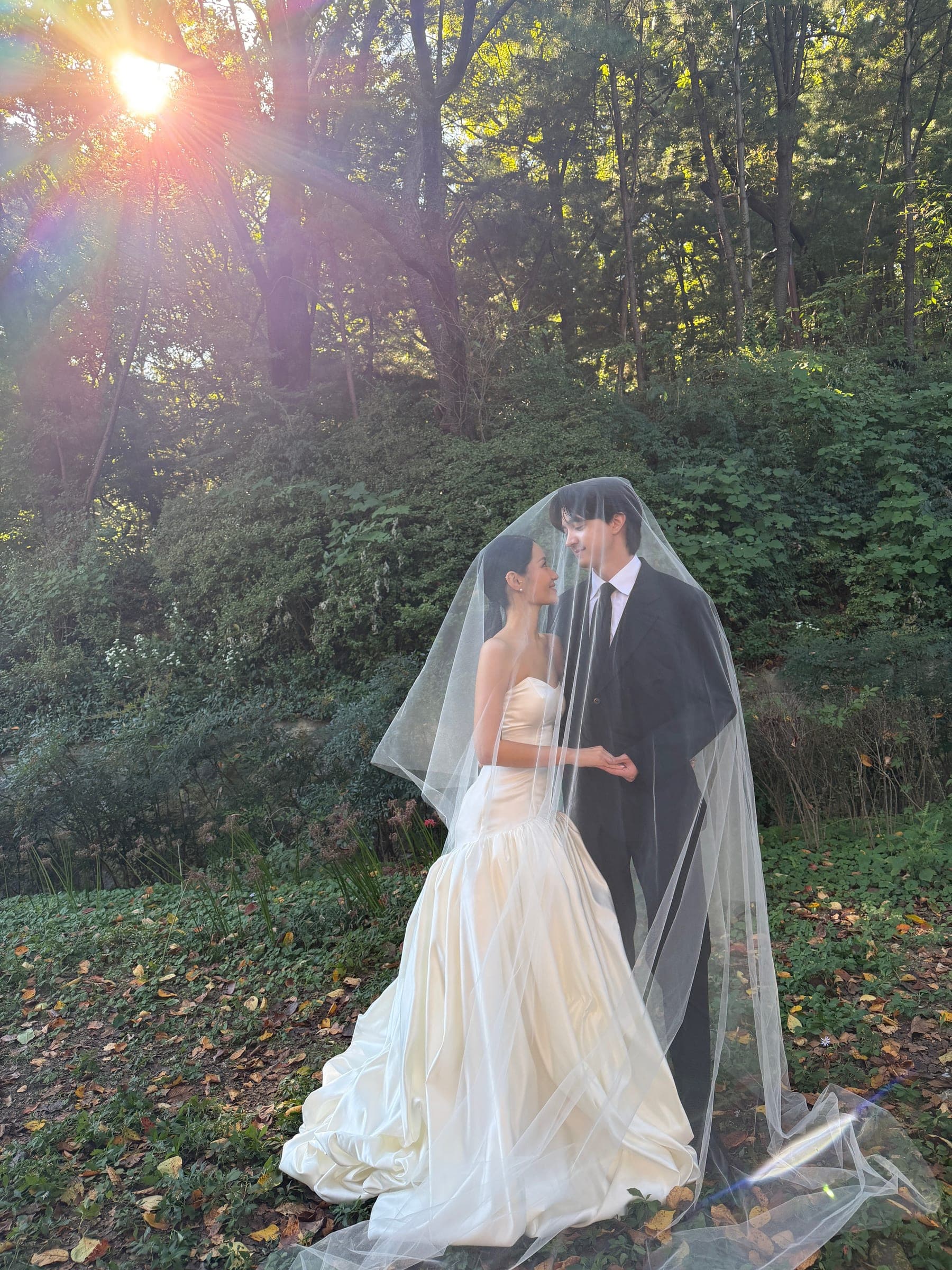 Chaewon and Brandon smiling at each other beneath a bridal veil, with sunlight streaming through the trees