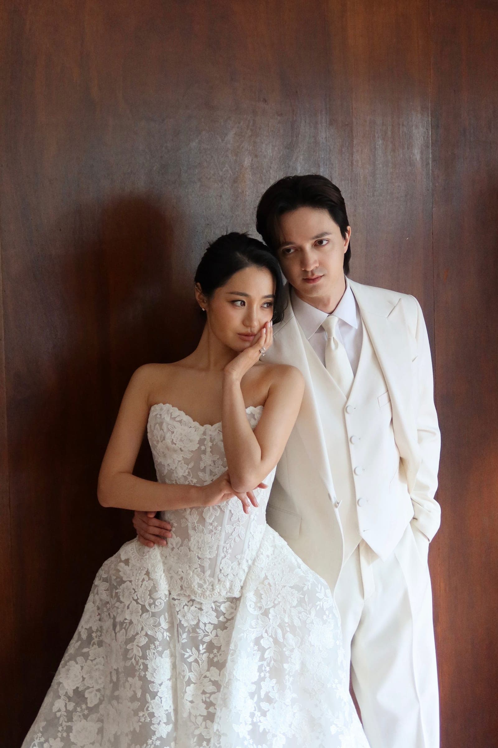 Chaewon in a lace gown and Brandon in an ivory suit, leaning together against a wooden wall